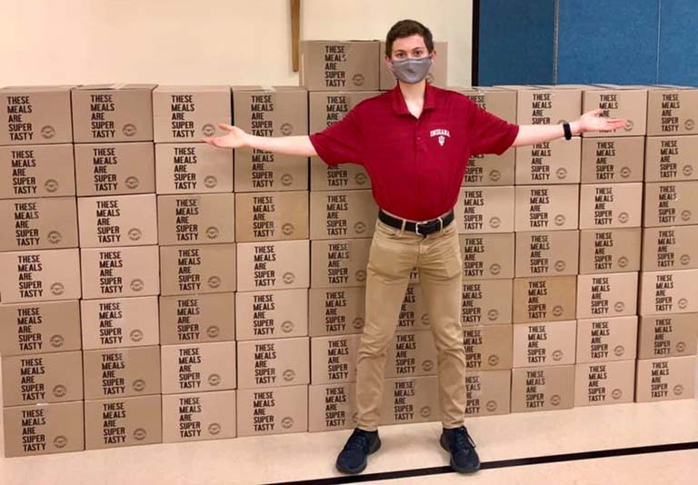 Matthew Ross poses in front of a wall of boxes containing meals prepared for children in the community.