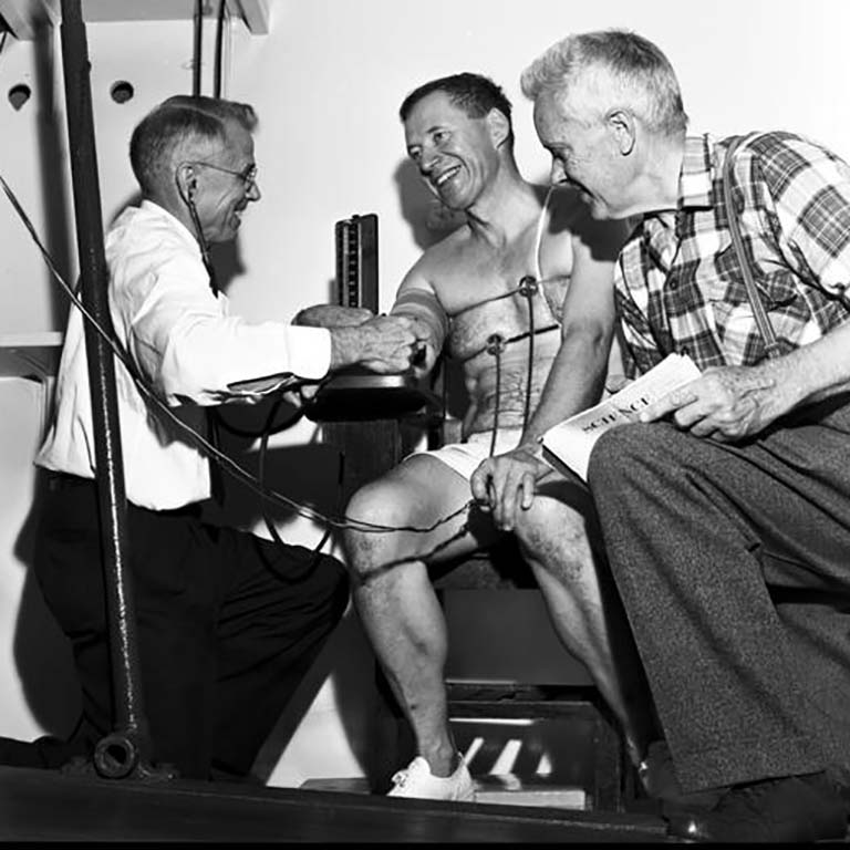 This image appears on page 27 of the December 1961 issue of the Indiana Alumni Magazine. The caption reads, in full: "Don Lash, famous as a runner for the University in the 1930's and now an Indianapolis FBI man, runs again on the treadmill in the physiology laboratory. Mr. Lash volunteered to participate in research being conducted by David B. Dill (right), research scholar, and Sid Robinson (left) head of the anatomy and physiology department, both former athletes also. Drs. Dill and Robinson are duplicating tests made many years ago on athletes, including themselves, to provide data on changes with age. Mr. Lash at the height of his athletic career cooperated with Robinson n extensive research on the physiological effects of exercise." Photo courtesy of IU Archives (Image #P0047097)