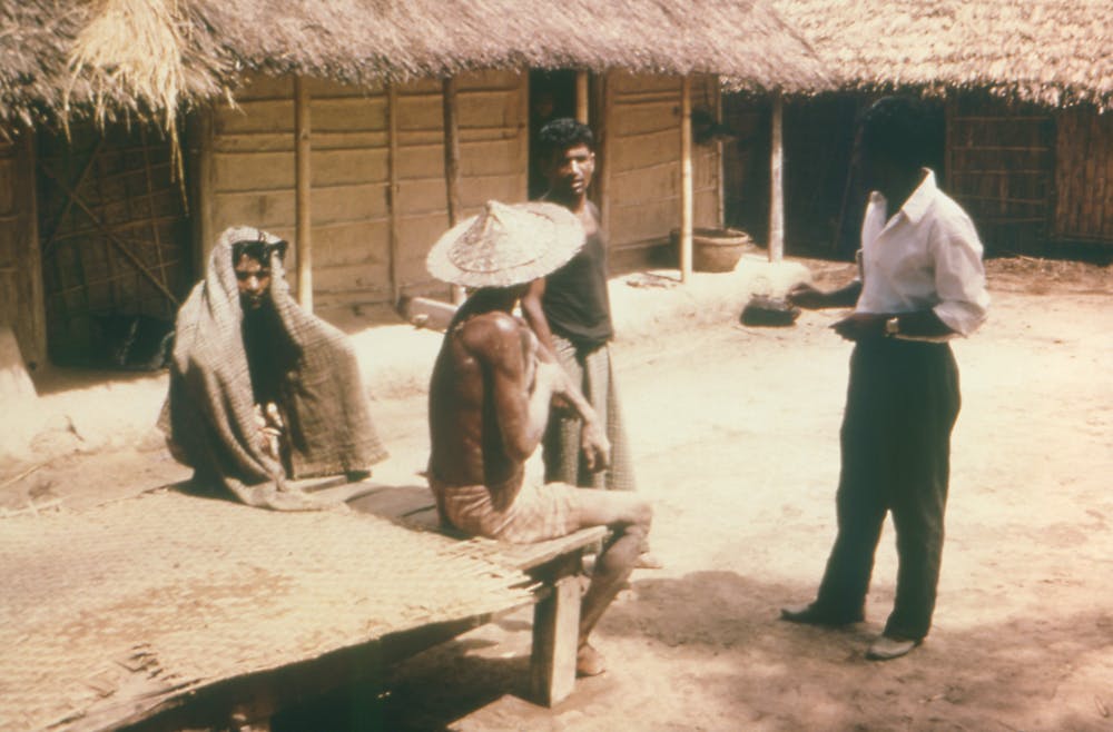 In 1977, a health care field worker conducts a house-to-house search for possible smallpox-infected inhabitants. He speaks with three men outside of a thatch-roofed house.