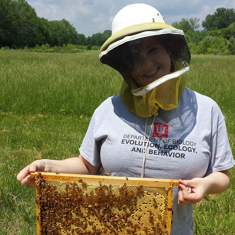 Delaney Miller wears a protective helmet and netting while holding up a honeycomb frame containing wax and bees.