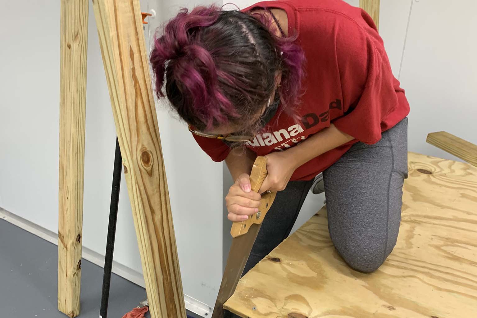 Victoria Lydick, a woman, kneels one knee on a plywood shelf as she bends over and cuts a strip from the end of the shelf with a hand saw.