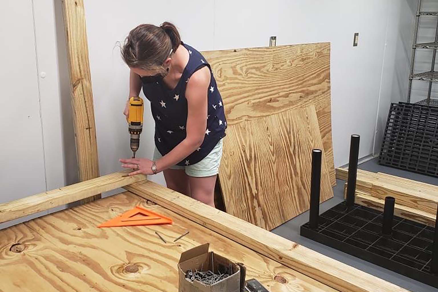 Julia van Kessel, a woman, uses an electric drill to secure the ends of two boards coming together at a right angle as she builds large plywood shelves.