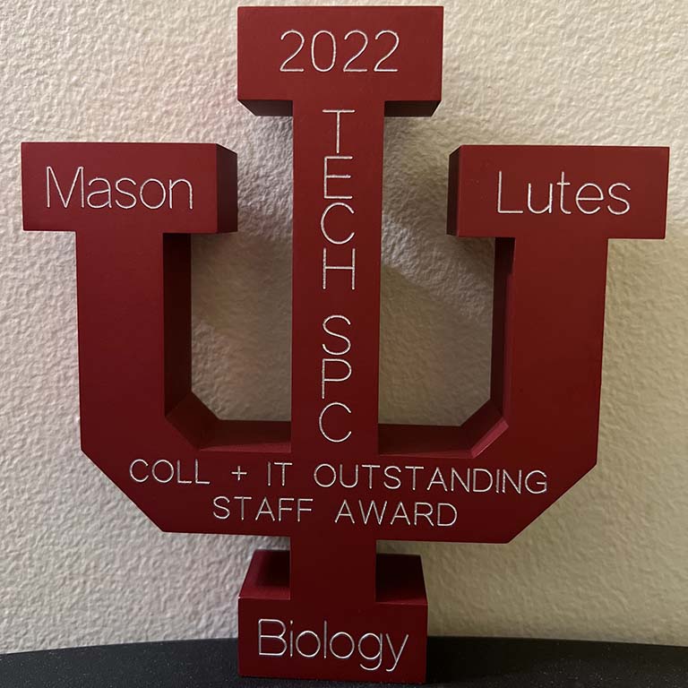 The front of the award medal received by Mason Lutes, recipient of the IU College of Arts and Sciences Outstanding IT Staff Award.  The medal is a red, IU-trident-shaped aluminum medal with the name of the award, Mason's name and title (tech specialist), "Biology" (Mason's department), and 2022 are engraved on the front side.