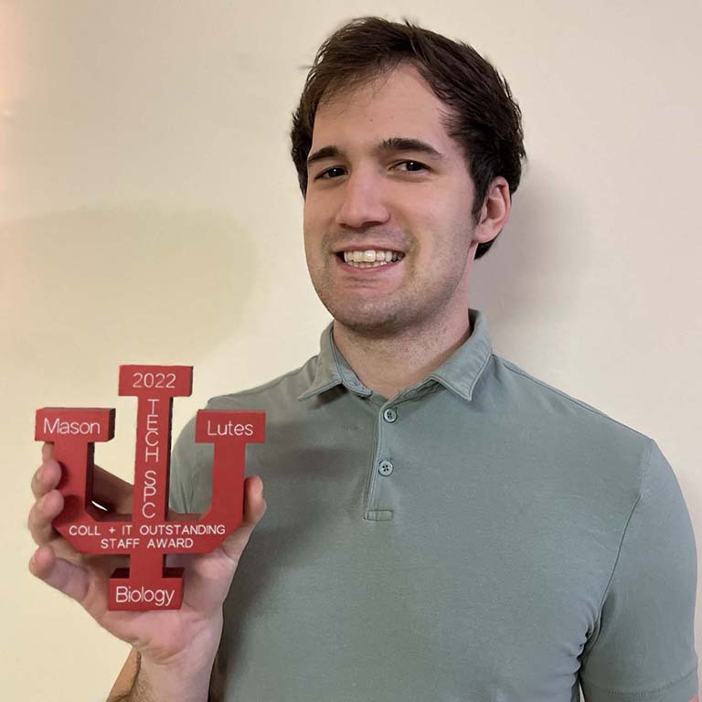 Mason Lutes, recipient of the IU College of Arts and Sciences Outstanding IT Staff Award, holds up the award medal he received.  The medal is a red, IU-trident-shaped aluminum medal with the name of the award, Mason's name and title (tech specialist), "Biology" (Mason's department), and 2022 engraved on the front side.