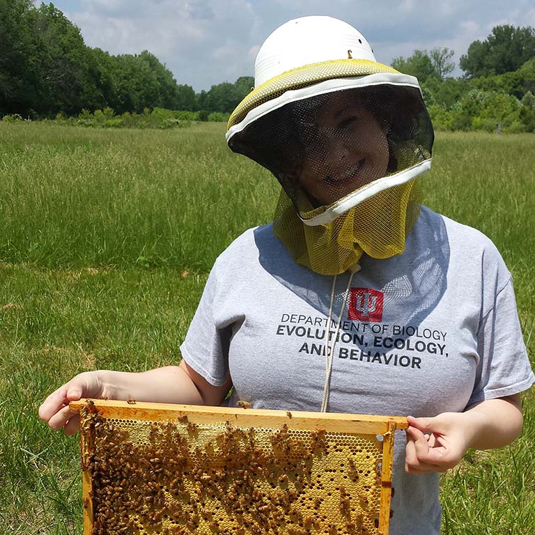 Delaney Miller, wearing a beekeeper's hood, holds an active honeycomb frame from a hive box.