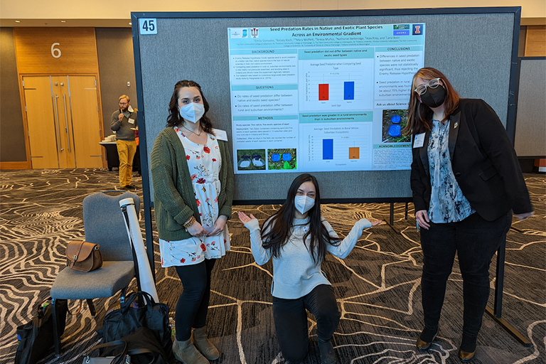 Three young women pose next to their research project poster "Seed Predation Rates in Native and Exotic Plant Species Across an Environmental Gradient."