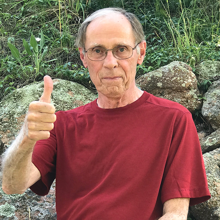 Norm Pace gives a thumbs-up while sitting on some rocks on a hillside.