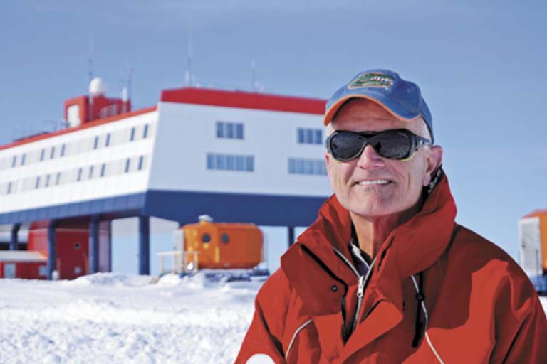 Rob Ferl on a research trip doing plant stress tests in Antarctica. Ferl poses for photo in front of a long building.  Blue sky and lots of snow!