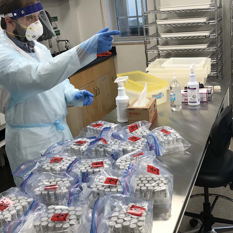 Lab assistant D.J. Ottman (dressed in protective clothing, gloves, face mask, and face shield) prepares to process the first set of samples. The vials of saliva that arrive at the lab in clear biohazard bags sit in front of him on a table in the testing facility.
