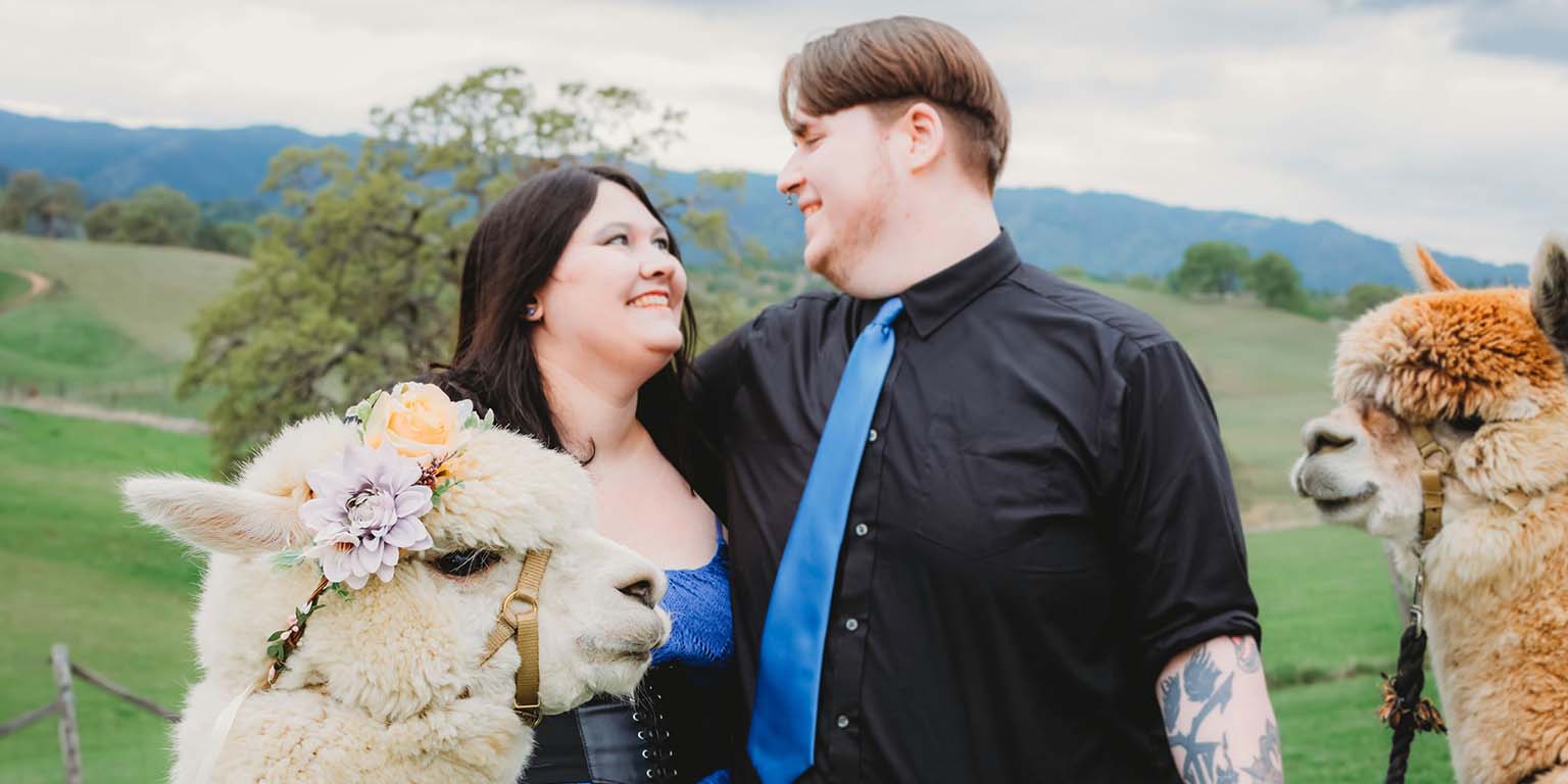 Gabrielle Sell and her husband looking lovingly at each other while standing with two alpacas in a pasture.