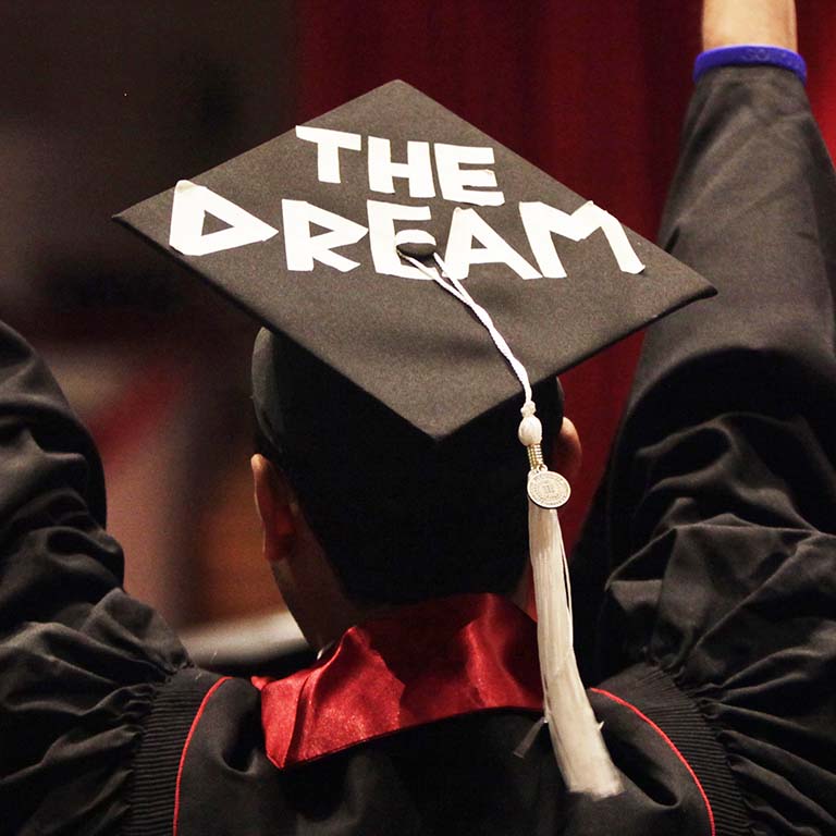 A graduating student wears a cap and tassel with large white letters:  THE DREAM.