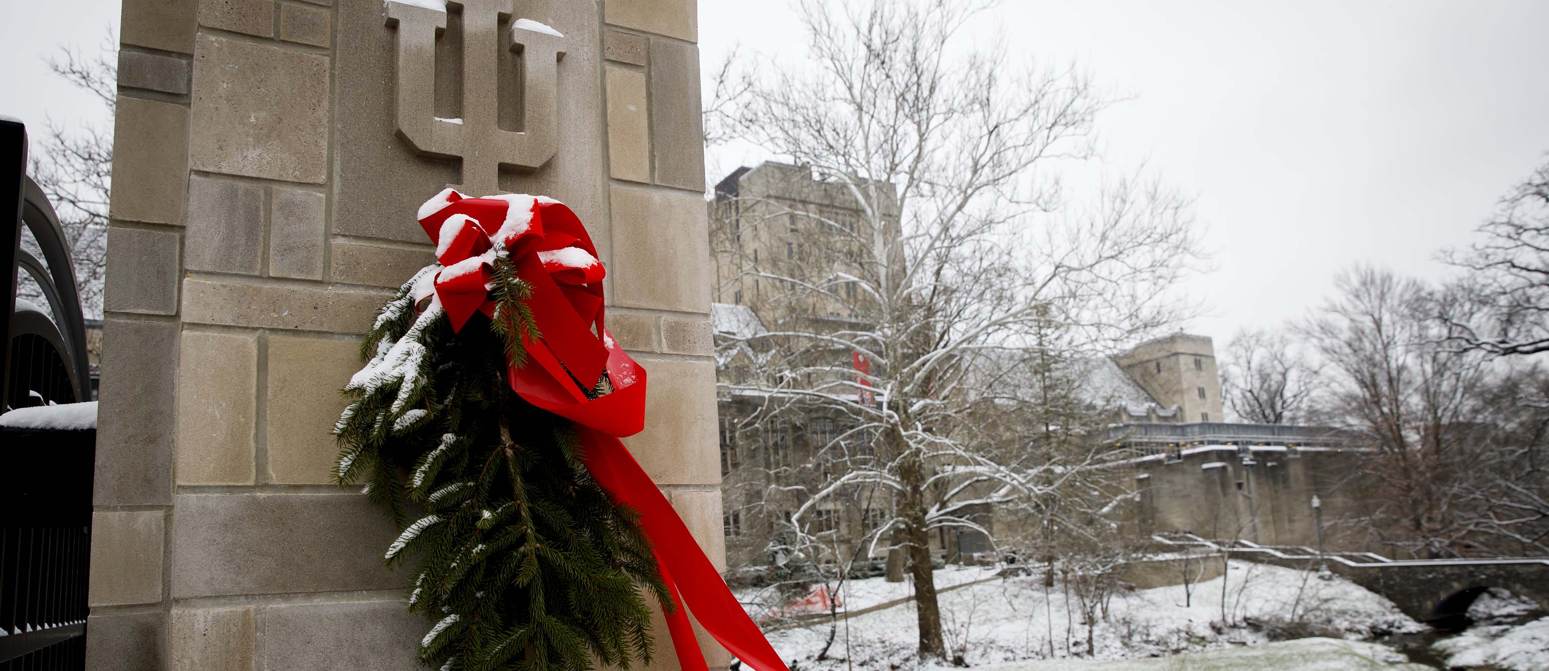 Pine branches bundled with a large red bow at the top hang just below a carved IU trident in a limestone gate post outside the Indiana Memorial Union. The IMU and Campus River are seen in the background. Snow covers the ground. There is snow on the decoration as well.