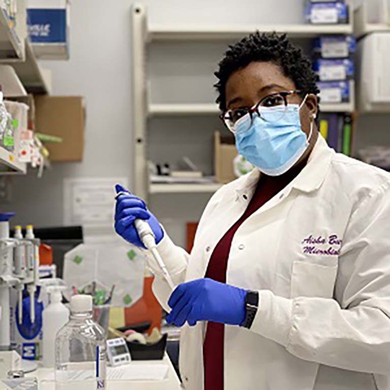 Aisha Burton, dressed in white lab coat and wearing blue gloves and face mask, holds a pipette while working in the lab.