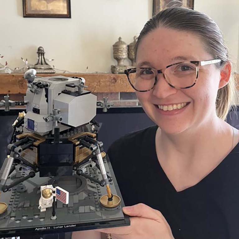 Brittany Herrin--a young, white woman with pulled-back brown hair and wearing glasses, holds a replica of the iconic NASA Apollo 11 Eagle Lunar Lander.