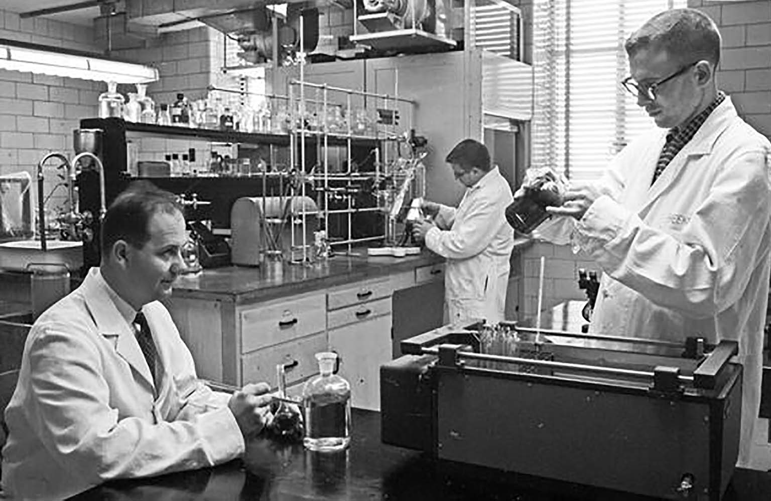 Frank Zeller (left) sitting at a table in his lab in May 1958. Two young men (students?) are working at benches in the lab.