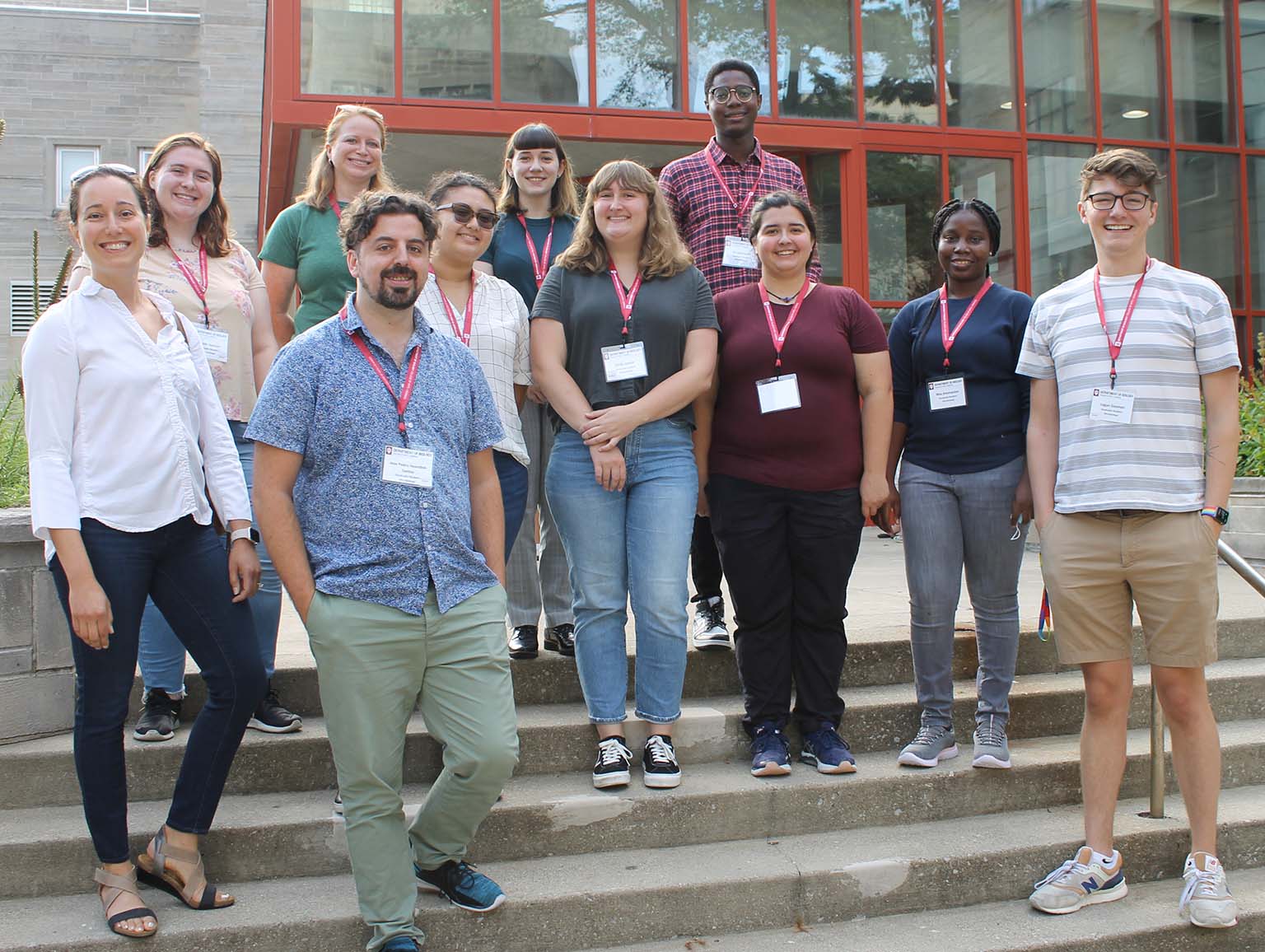 Logan Geyman (right) stands with nine other young women and men and Professor Irene Newton (left) on the steps outside of the Biology Building atrium while posing for a group photo of the incoming Microbiology graduate students during orientation.