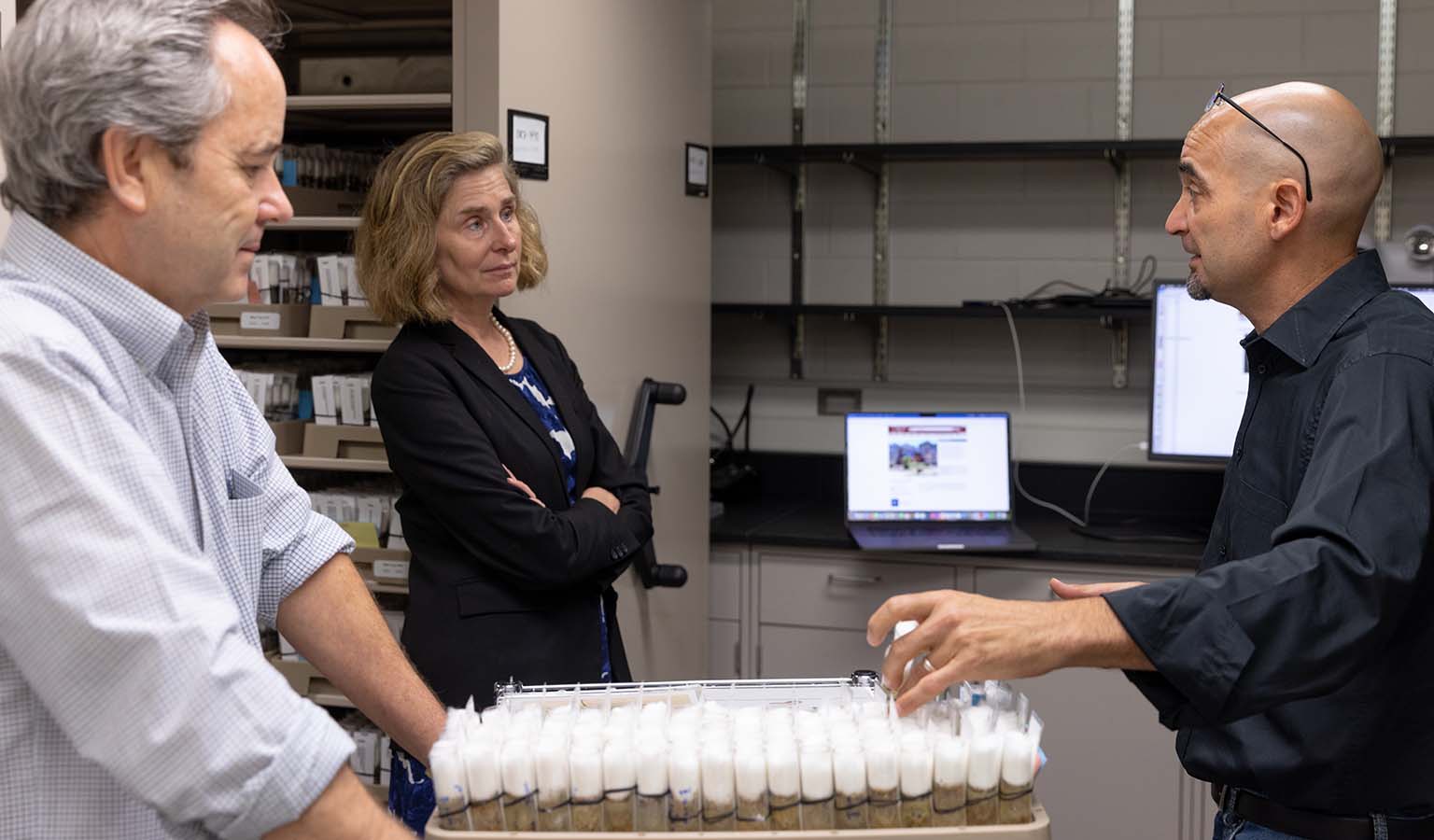 Kevin Cook (left) looks on as Andy Zelhof (right) talks to Pamela Whitten. Cook has brought out a cart with a large tray of cotton-topped vials with living fly cultures.